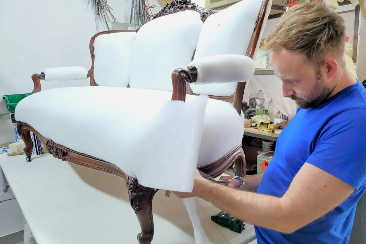 Restauro divani e poltrone: il tappezziere ritaglia l'ovatta sulla nuova imbottitura di un divanetto a tre schienali